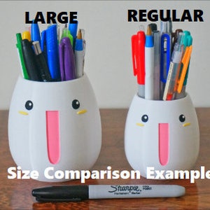 May include: Size comparison of two white ceramic pencil holders with cute faces. The larger holder is labeled "LARGE" and the smaller holder is labeled "REGULAR". Both holders are filled with various colored pens and pencils.