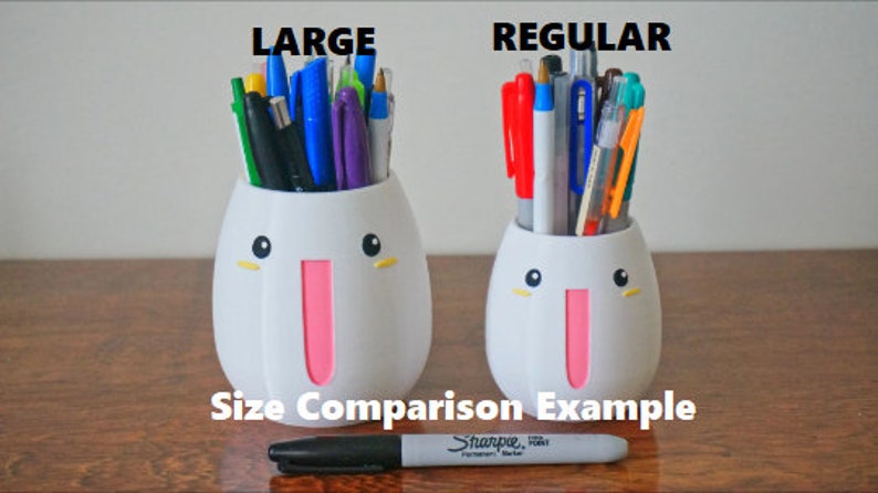 May include: Size comparison of two white ceramic pencil holders with cute faces. The larger holder is labeled "LARGE" and the smaller holder is labeled "REGULAR". Both holders are filled with various colored pens and pencils.