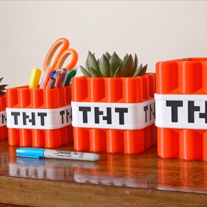 May include: Four orange Minecraft TNT-themed containers with white labels reading "TNT". The containers are arranged in a row from smallest to largest. The smallest container holds a small succulent plant, the next holds pens and scissors, the third holds a larger succulent plant, and the largest container is empty.