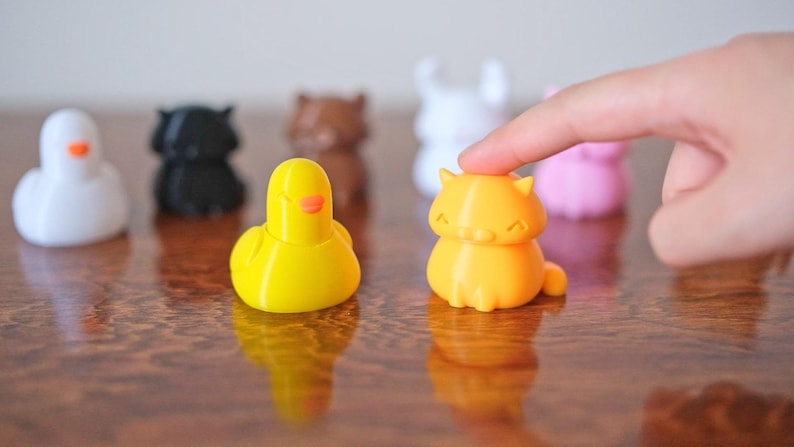 Peut inclure: Une collection de petites figurines d'animaux en 3D imprim&eacute;es en couleur, comprenant un canard, un chat, un lapin, un ours et un cygne. Les figurines sont dispos&eacute;es sur une surface en bois.