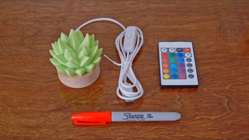 K&ouml;nnte beinhalten: Eine hellgr&uuml;ne LED-Lampe in Form einer Sukkulente mit Holzsockel, Fernbedienung und wei&szlig;em Kabel. Ein roter und grauer Sharpie-Feinmarker ist ebenfalls vorhanden.
