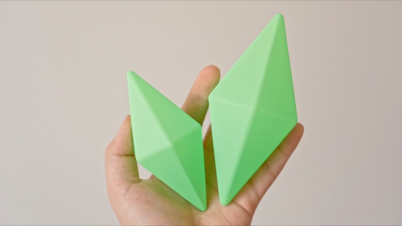 May include: Two green geometric shapes, resembling diamonds, are held in a hand. The shapes are made of a smooth, matte material.