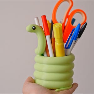 May include: A light green, snake-shaped desk organizer filled with pens, markers, and scissors. The organizer is held in a hand and has a coiled body design. The scissors are orange, and the pens and markers are various colors.