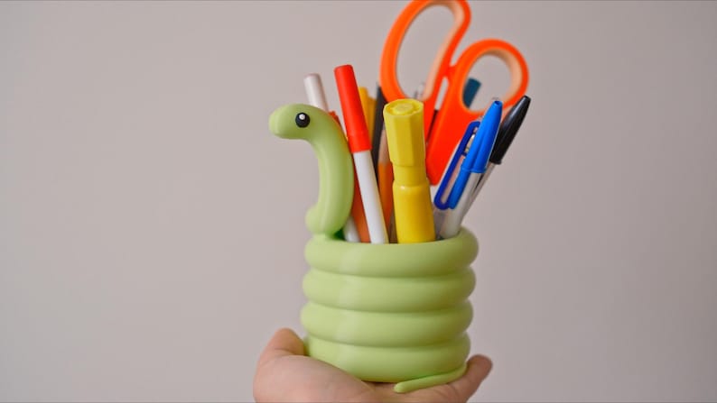 May include: A light green, snake-shaped desk organizer filled with pens, markers, and scissors. The organizer is held in a hand and has a coiled body design. The scissors are orange, and the pens and markers are various colors.