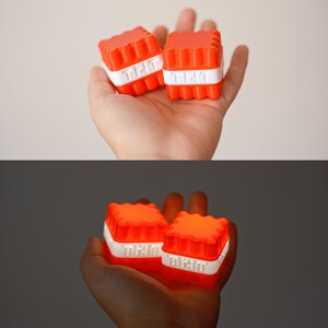 Op de afbeelding: Twee kleine, oranje, vierkante lichtgevende speelgoedjes met witte tekst die op elk "Tlit" staat. De speelgoedjes worden in de hand van een persoon gehouden.