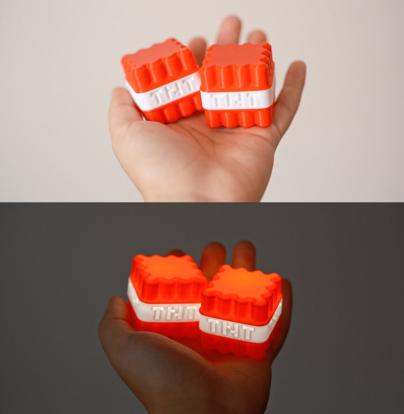 Op de afbeelding: Twee kleine, oranje, vierkante lichtgevende speelgoedjes met witte tekst die op elk "Tlit" staat. De speelgoedjes worden in de hand van een persoon gehouden.