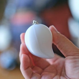 May include: A white, mango-shaped keychain with a silver-colored metal loop. The smooth, glossy surface reflects light, creating highlights. The keychain is held in a hand, with a blurred background.