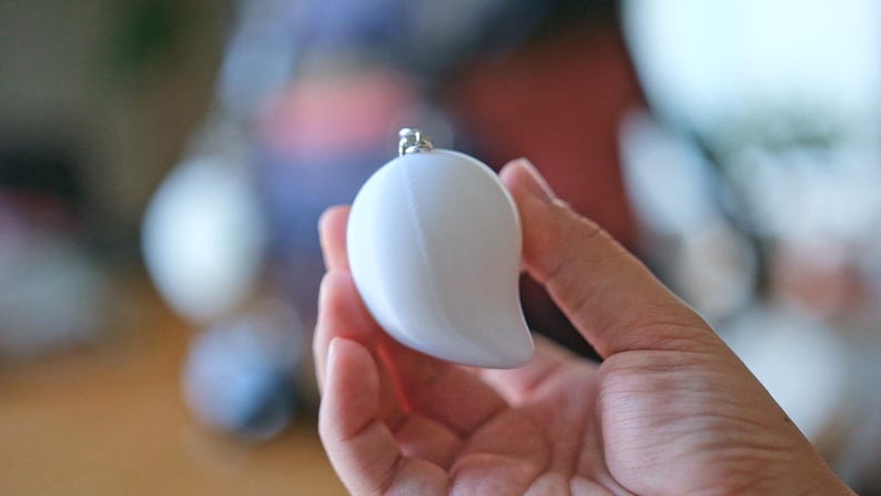 May include: A white, mango-shaped keychain with a silver-colored metal loop. The smooth, glossy surface reflects light, creating highlights. The keychain is held in a hand, with a blurred background.