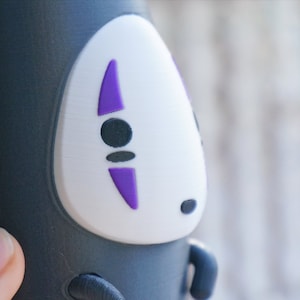 May include: A black and white No-Face figure from the animated film Spirited Away. The figure has a white face with purple accents and black eyes. The figure is a cylindrical shape.