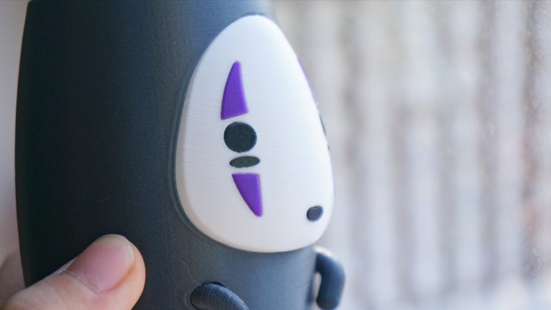 May include: A black and white No-Face figure from the animated film Spirited Away. The figure has a white face with purple accents and black eyes. The figure is a cylindrical shape.