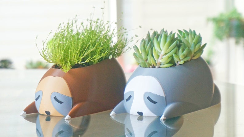 May include: Two ceramic sloth planters, one brown and one gray, with green plants growing out of the top. The sloths have closed eyes and are lying down.
