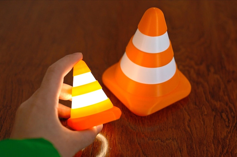 Op de afbeelding: Twee oranje en witte verkeerskegels, een klein en een groot. De kleinere kegel is verlicht en wordt in een hand gehouden.