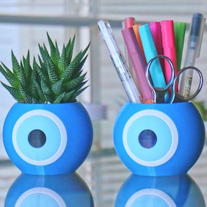 Puede incluir: Dos macetas de cerámica azul con un diseño de ojo turco blanco. Una maceta tiene una pequeña planta suculenta, la otra tiene una variedad de bolígrafos y tijeras.