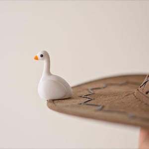 May include: A small, white goose figurine with an orange beak sits on the brim of a brown cowboy hat. The hat has decorative stitching and a classic western style. The background is a soft, neutral color.