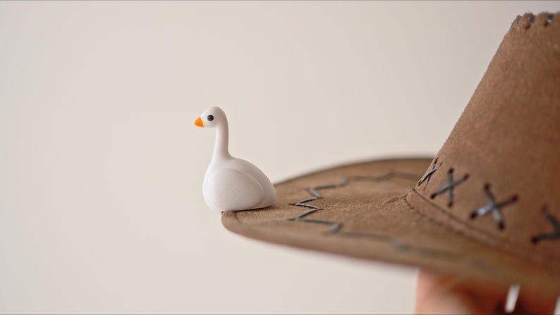 May include: A small, white goose figurine with an orange beak sits on the brim of a brown cowboy hat. The hat has decorative stitching and a classic western style. The background is a soft, neutral color.