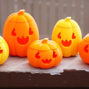 May include: Four orange and yellow plastic jack-o'-lantern candy holders with carved faces. The holders are different sizes and are arranged on a wooden surface.