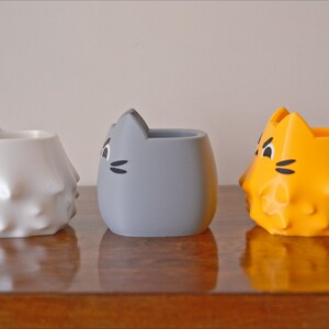 May include: Three ceramic cat-shaped planters, one white, one gray, and one orange. The planters have a textured surface and a cute cat face design.
