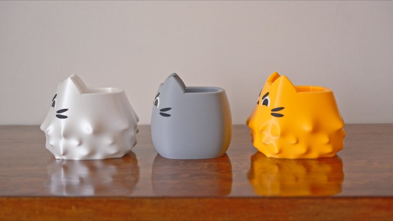 May include: Three ceramic cat-shaped planters, one white, one gray, and one orange. The planters have a textured surface and a cute cat face design.