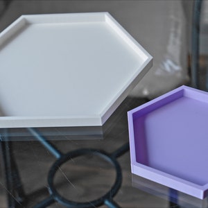 May include: Two hexagonal trays, one white and one lavender, are displayed on a glass surface. The trays are made of a smooth, solid material and have a raised edge.
