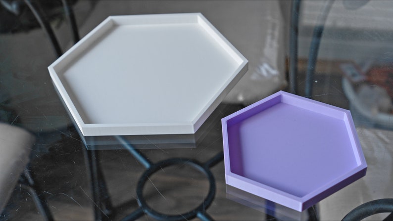 May include: Two hexagonal trays, one white and one lavender, are displayed on a glass surface. The trays are made of a smooth, solid material and have a raised edge.
