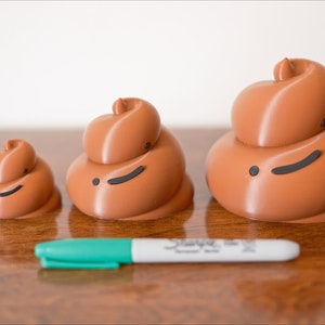 May include: Three brown ceramic figurines shaped like a pile of poop, with black eyes and a mouth, arranged in order of size from smallest to largest on a wooden surface. A green marker is in front of the figurines.