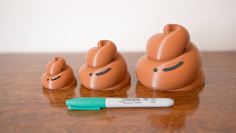 May include: Three brown ceramic figurines shaped like a pile of poop, with black eyes and a mouth, arranged in order of size from smallest to largest on a wooden surface. A green marker is in front of the figurines.