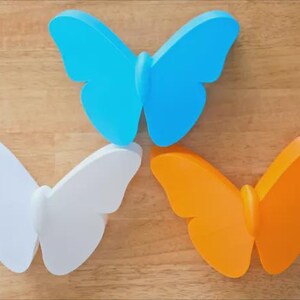 May include: Three colorful butterfly decorations. One is light blue, one is white, and the other is orange. The butterflies are arranged on a wooden surface.