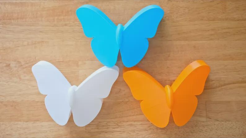 May include: Three colorful butterfly decorations. One is light blue, one is white, and the other is orange. The butterflies are arranged on a wooden surface.