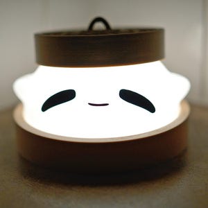 Puede incluir: Una luz blanca con una cara sonriente, que se asemeja a una bola de masa, se ilumina dentro de un recipiente de madera. El recipiente tiene una tapa con un dise&ntilde;o de bamb&uacute;.