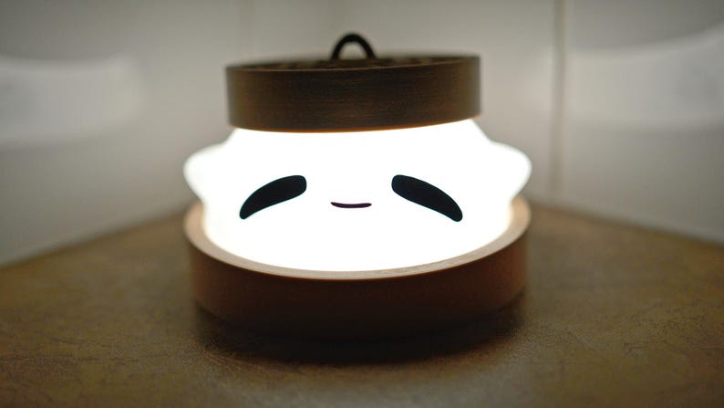 Puede incluir: Una luz blanca con una cara sonriente, que se asemeja a una bola de masa, se ilumina dentro de un recipiente de madera. El recipiente tiene una tapa con un dise&ntilde;o de bamb&uacute;.