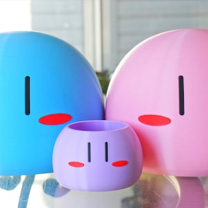 May include: Three dango figurines, one blue, one pink, and one purple. Each figurine has a simple face with two black eyes and two red cheeks.