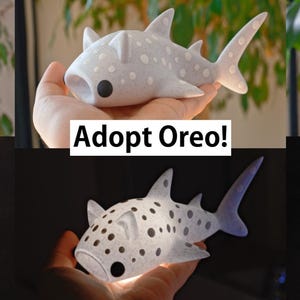 May include: Three white plush shark toys with black spots. Each shark has a different name: Duck, Oreo, and Sesame. The sharks are being held in a person's hand. The text "Adopt Duck!" is below the first shark, "Adopt Oreo!" is below the second shark, and "Adopt Sesame!" is below the third shark.