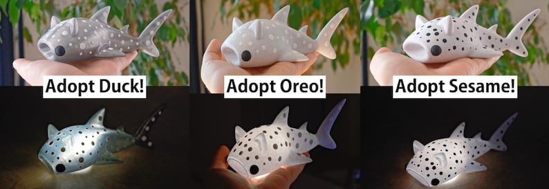 May include: Three white plush shark toys with black spots. Each shark has a different name: Duck, Oreo, and Sesame. The sharks are being held in a person's hand. The text "Adopt Duck!" is below the first shark, "Adopt Oreo!" is below the second shark, and "Adopt Sesame!" is below the third shark.