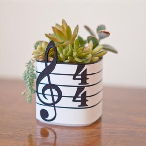 May include: A white ceramic planter with a musical theme, featuring a black treble clef and the numbers 4/4. The planter is filled with a variety of succulents in shades of green and yellow, creating a decorative accent.