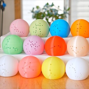 May include: A collection of twelve colorful, spherical objects, each with a constellation pattern of small holes. The spheres are arranged on a white surface, with colors including pink, green, blue, orange, and yellow. The objects are likely decorative or functional.