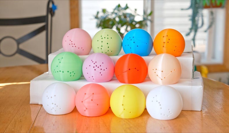 May include: A collection of twelve colorful, spherical objects, each with a constellation pattern of small holes. The spheres are arranged on a white surface, with colors including pink, green, blue, orange, and yellow. The objects are likely decorative or functional.