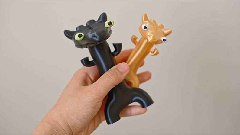 May include: Two dragon-shaped bottle openers. One is black with green eyes, the other is gold with blue eyes. The openers are held in a hand against a neutral background. The dragon openers have a tail and small arms.
