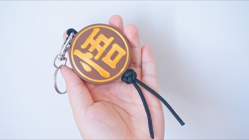Puede incluir: Un llavero circular marr&oacute;n con un car&aacute;cter chino dorado "道" (Dao) en &eacute;l. El llavero tiene un cord&oacute;n negro con un nudo en el extremo.
