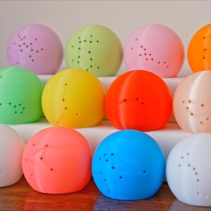 May include: A collection of twelve colorful, spherical objects, each with a constellation pattern. The spheres are arranged in three rows, showcasing a variety of colors including pink, green, yellow, orange, and blue. The objects are displayed on a white surface.