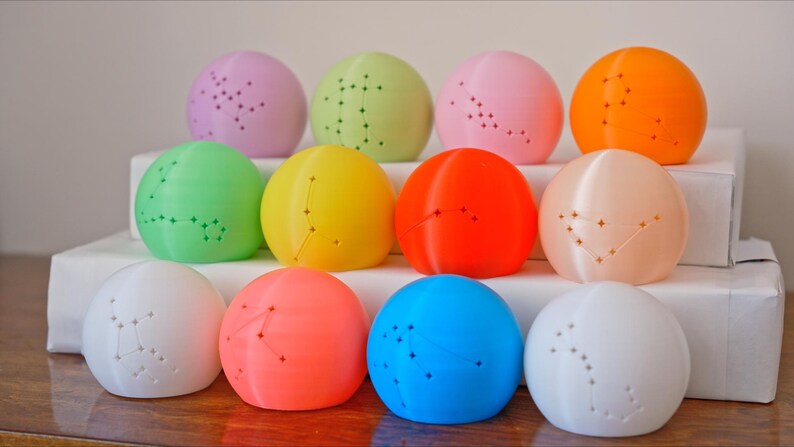 May include: A collection of twelve colorful, spherical objects, each with a constellation pattern. The spheres are arranged in three rows, showcasing a variety of colors including pink, green, yellow, orange, and blue. The objects are displayed on a white surface.