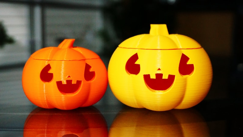 May include: Two Halloween jack-o'-lantern decorations, one orange and one yellow, with carved smiling faces. The smaller orange pumpkin is on the left and the larger yellow pumpkin is on the right.