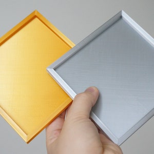K&ouml;nnte beinhalten: Zwei quadratische Tabletts, eines golden und eines silbern, mit einem erh&ouml;hten Rand um den Umfang. Die Tabletts sind aus einem strukturierten Material gefertigt.