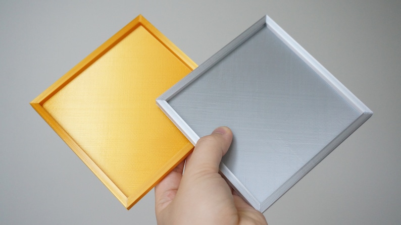 K&ouml;nnte beinhalten: Zwei quadratische Tabletts, eines golden und eines silbern, mit einem erh&ouml;hten Rand um den Umfang. Die Tabletts sind aus einem strukturierten Material gefertigt.