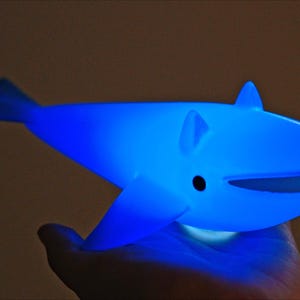 May include: A glowing blue whale-shaped night light. The whale has fins, a tail, and a smiling mouth. The light is held in a hand, and the background is a dark brown.