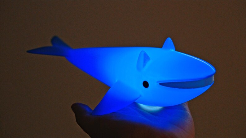 May include: A glowing blue whale-shaped night light. The whale has fins, a tail, and a smiling mouth. The light is held in a hand, and the background is a dark brown.