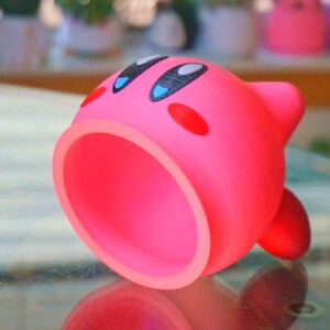 May include: A pink 3D printed character with a large open mouth, black eyes, and red cheeks. The character is lying on its side on a glass surface.