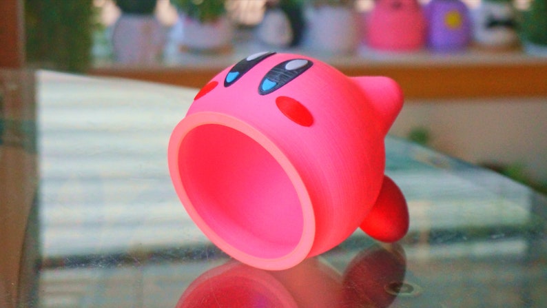May include: A pink 3D printed character with a large open mouth, black eyes, and red cheeks. The character is lying on its side on a glass surface.