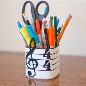May include: A white pen holder decorated with a musical staff and a treble clef, holding various pens, pencils, and scissors. The pen holder has the numbers 4/4 printed on it. The items inside are of various colors.