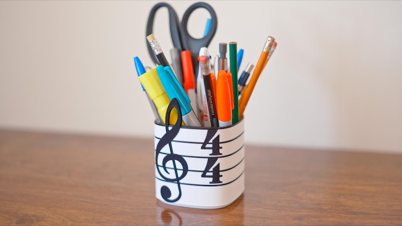 May include: A white pen holder decorated with a musical staff and a treble clef, holding various pens, pencils, and scissors. The pen holder has the numbers 4/4 printed on it. The items inside are of various colors.
