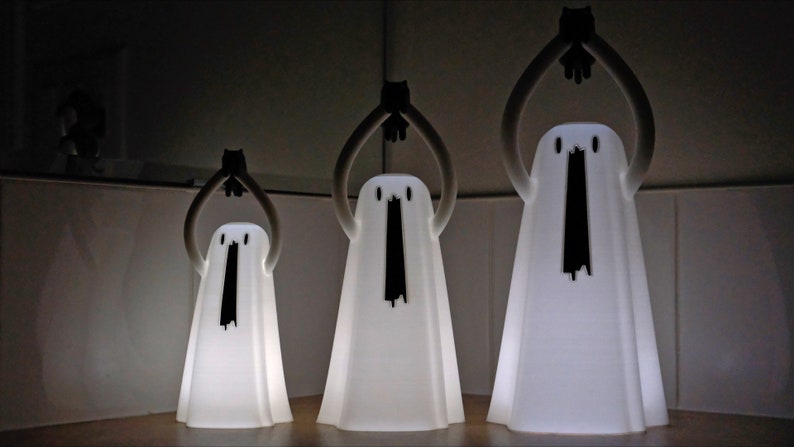 Peut inclure: Trois lampes blanches en forme de fant&ocirc;me avec des yeux et une bouche noirs, pos&eacute;es sur une surface en bois. Les fant&ocirc;mes ont les bras lev&eacute;s au-dessus de leur t&ecirc;te.